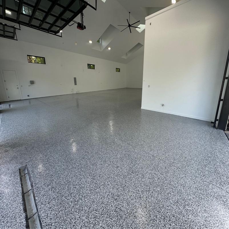 Spacious, modern garage with high ceilings, skylights, and polished speckled floor. Walls are white with minimal decor. Large fan is mounted overhead.