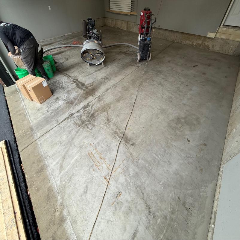 A person works in a garage on concrete with machinery, preparing the surface. Several green buckets and boxes are scattered around.