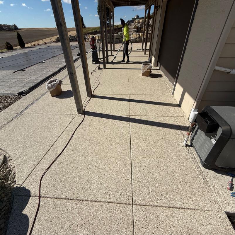A person works on a sunny patio with a power washer, surrounded by barren fields and garden features, under an elevated porch.