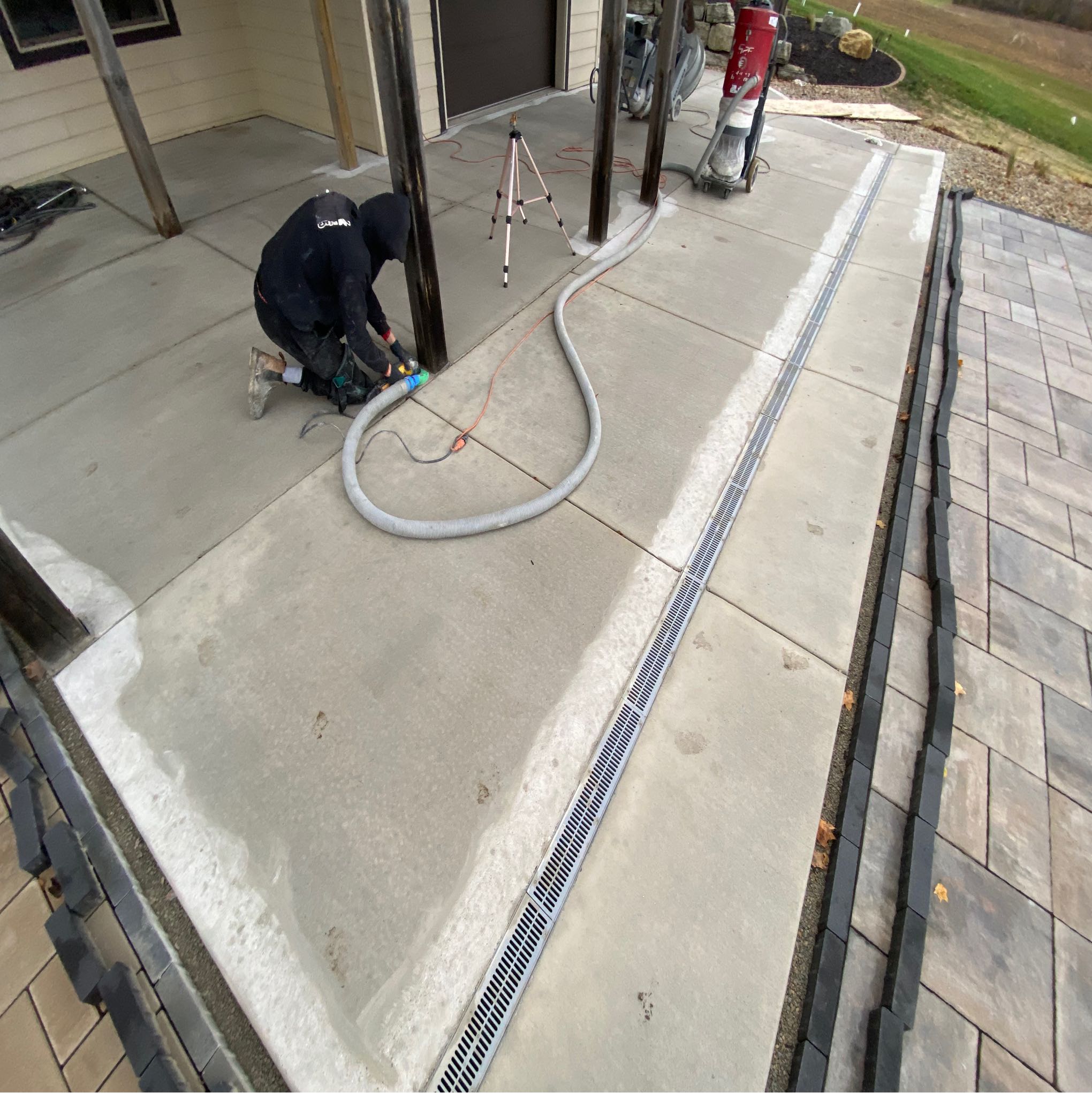 A person wearing a hood repairs a concrete patio, with tools and a vacuum nearby, at a residential outdoor area.