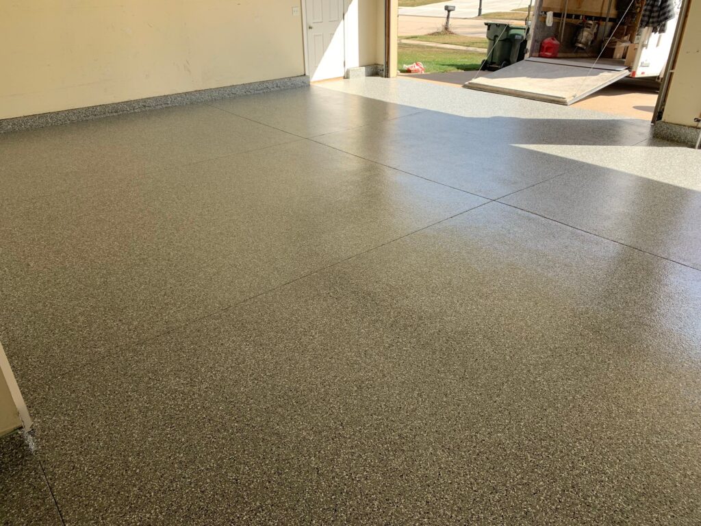 A clean, empty garage with polished concrete flooring, open to a sunlit driveway and scattered tools in the background, no people present.