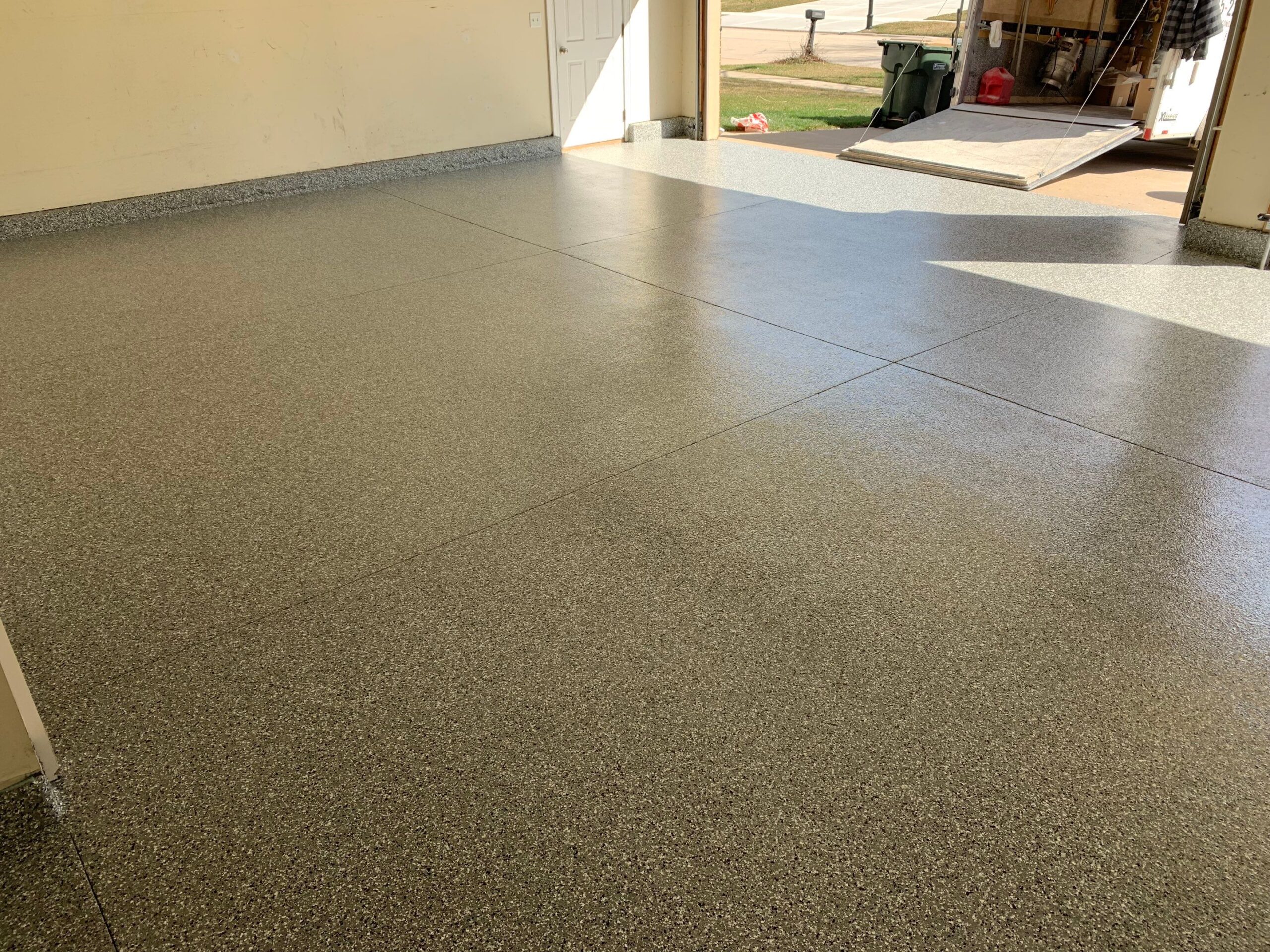 Empty garage with newly finished epoxy floor, open to driveway. Sunlight casts shadows inside, with tools and equipment visible outside.
