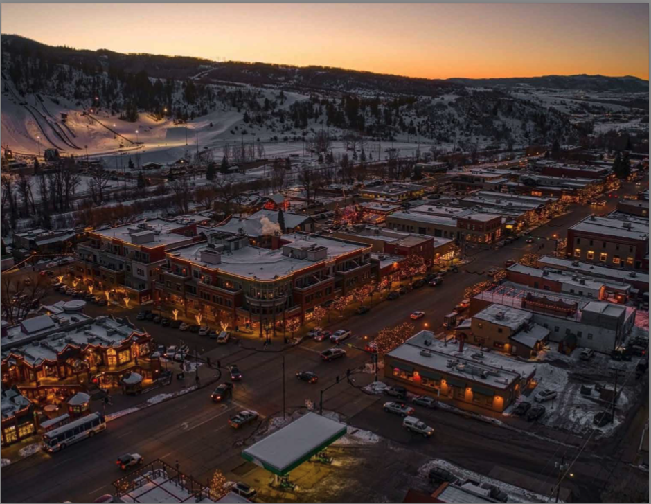 Snowy town at dusk with festive lights, surrounded by hills. Illuminated streets, buildings, and a gas station create a cozy winter atmosphere.